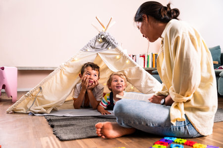 Asian family at home - asian mother reads aloud to two delighted children inside a cozy teepee at...