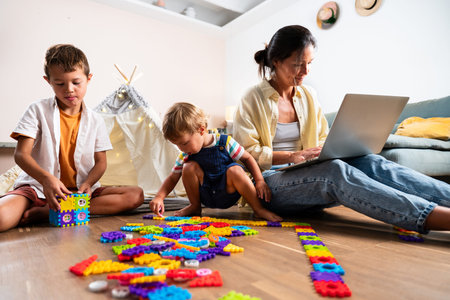 Asian family at home - asian mother multitasking, working from home on a laptop while her two young...