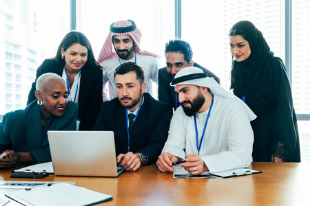 Diverse international business professionals gathered around a laptop in a modern corporate office, collaborating and discussing strategy during a productive team meeting