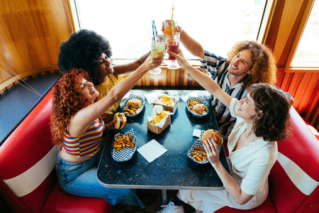 Multiethnic group of young happy friends bonding at vintage diner burger house restaurant - diverse group of young friends enjoying toasts and burgers in a retro american diner
