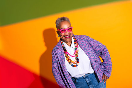 Black senior woman wearing modern clothing portrait on background - senior black woman wearing pink sunglasses, colorful necklaces, and a plaid jacket, expressing joy against a bright background