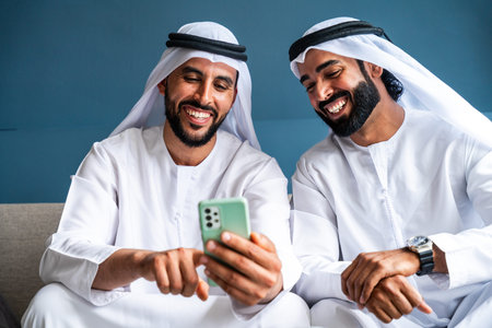 Two smiling adult arab men in traditional clothing. Ghutra and kandura. Are sitting on a sofa. Happily sharing and interacting with content on a smartphone. Representing connection. Modern technology
