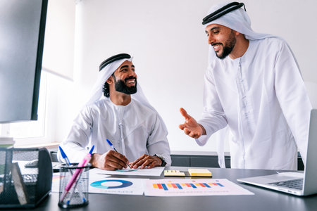Arab men in traditional clothing collaborating and smiling while reviewing charts and working on financial reports, fostering a positive business environment