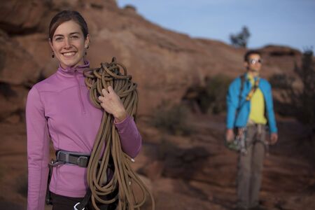 An attractive young woman is smiling and holding a climbing rope as her partner stands in the background. Horizontal shot.の写真素材
