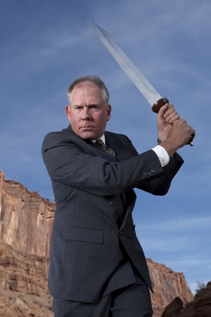 A businessman is holding a sword in an attack posture in a desert setting. Horizontal shot.の写真素材