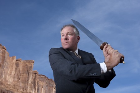 A businessman is holding a sword in an attack posture in a desert setting. Horizontal shot.の写真素材