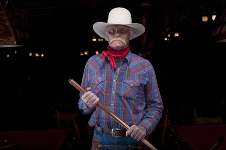 A senior man wearing a hat and western attire is standing in a darkened room holding a pool cue. He has an angry facial expression. Horizontal shot.の写真素材