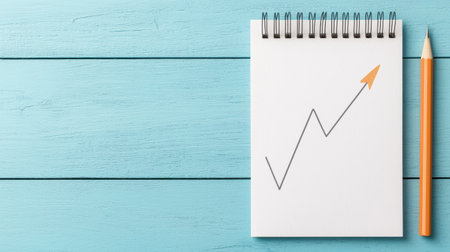 A notepad with a growth chart drawing and an orange pencil against a light blue wooden background.の素材