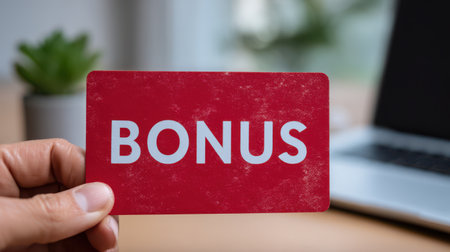 Hand Holding Red Bonus Card in Modern Workplace with Laptop and Plant in Background, Symbolizing Rewards and Incentives for Employees or Customersの素材