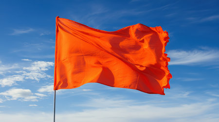 Bright orange flag waving against blue sky with white clouds in a vibrant outdoor setting, symbolizing warning, caution, or attention in various contextsの素材