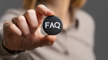 Hand Holding a Round FAQ Token Symbolizing Frequently Asked Questions in a Professional or Educational Setting with a Neutral Backgroundの素材
