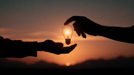 Hands Sharing a Light Bulb Against a Beautiful Sunset Sky Symbolizing Ideas, Inspiration, and the Power of Innovation in a Creative Environmentの素材