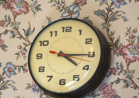 Old analog clock resting on wall with floral wallpaper.の写真素材