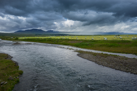 Iceland - Hundreds of hay bales behind river on green meadow with intense rain clouds and tractor while thunderstormの写真素材