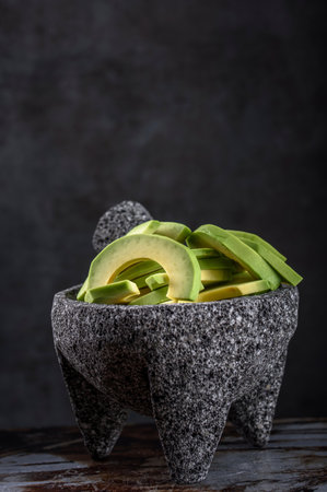 Avocado slices in stone mortar on wooden table. Dark background.