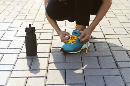 Young woman tying shoelaces on sneakers on a pavers. Standing next to a bottle of water. Exercise outdoorsの写真素材