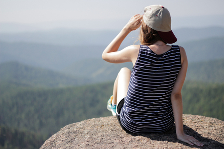 A young woman with a baseball cap on his head, sitting on a rock and looking at the horizon.の写真素材