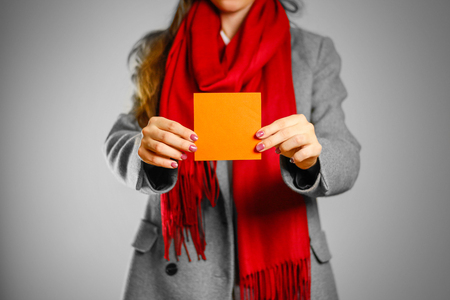 A girl in grey coat and red scarf holds a orange clean empty square sticker. Isolated on grey background.の写真素材