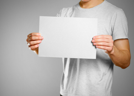 A man in a gray t-shirt keeps a grey clean blank sheet of A4. Isolated on grey background.の写真素材