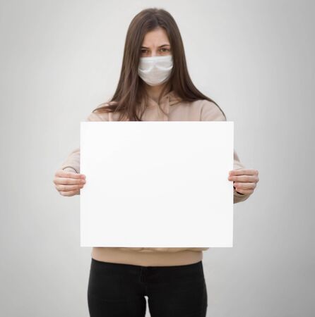 A girl in a medical mask holds an empty white sheet of paper. Close up. Isolated on a grey background.の写真素材