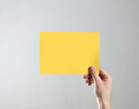 The hand holds an empty yellow sheet of paper. A clean flyer in your hands. For your text. Isolated on a gray background.の写真素材