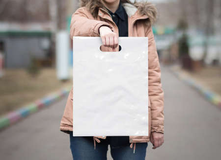 The girl is holding a mockup white plastic bag. A girl with a package on the street in the park.