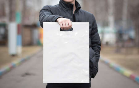 A man holds a mockup white plastic bag. A man with a package on the street in the park.