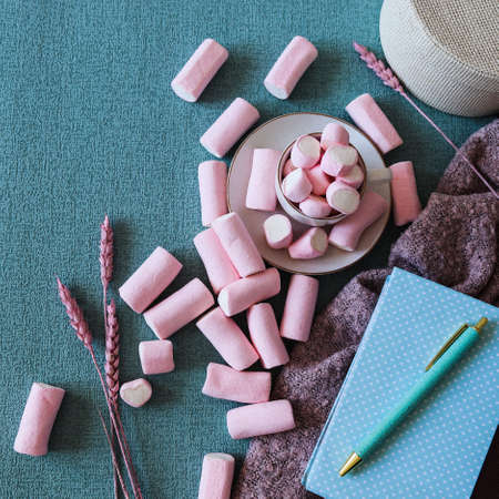 Sweet pink marshmallows on blue background. Blue desk with notebook and pen. A marshmallow is shaped like a heart. Along with dried pink flowers.の写真素材