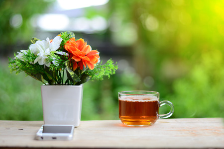 the white and orange  flowers in a white vase with smartphone and a cup of tea  on wooden table and orange light in morningの写真素材