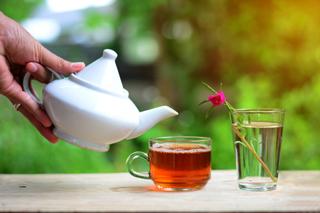 morning tea time , a hand holding a tea pot ,a cup of tea and glass vase with flower on wooden tableの写真素材