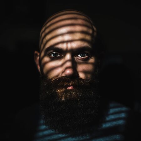 Close-up portrait of middle aged brutal bearded man with expressive eyes and striped shadows on his face with black backgroundの写真素材