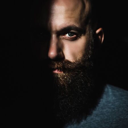 Close-up portrait of middle aged brutal bearded man with expressive eyes and shadow on half of his face with black backgroundの写真素材