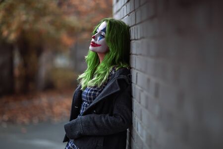 Portrait of a green haired girl in checkered dress with clown makeup on a brick wall background.