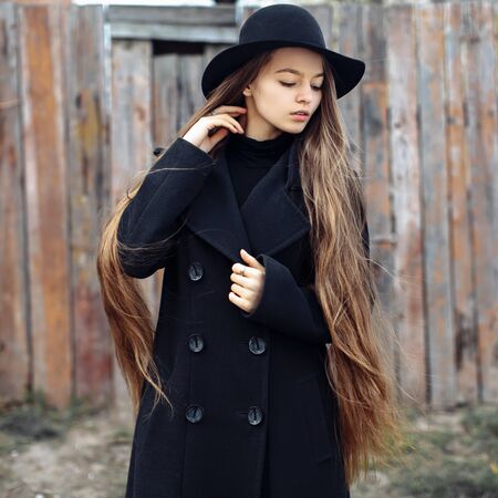 Young beautiful fashionable woman in black hat, with long hair posing on woody background. Female fashion, beauty concept. Outdoor.の写真素材