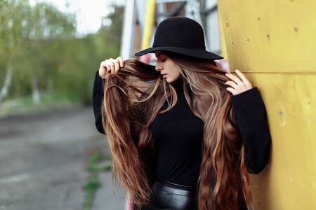 Young beautiful fashionable woman in black hat with long hair posing on blurry backgroundの写真素材