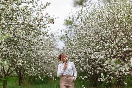 Beautiful young blonde woman in white shirt posing under apple tree in blossom in Spring gardenの写真素材