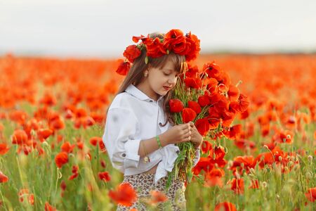Little cute girl in white dress playing field poppy wreath with a bouquet of poppies in her hands at summer sunsetの写真素材