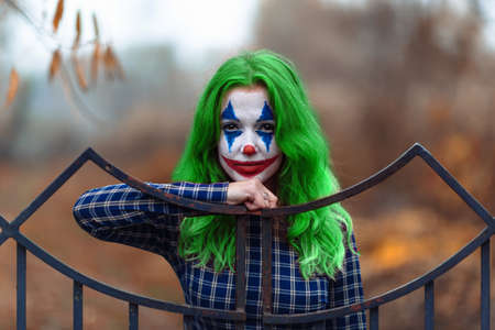 Close-up portrait of a greenhaired girl in chekered dress with joker makeup on a blurry brown...