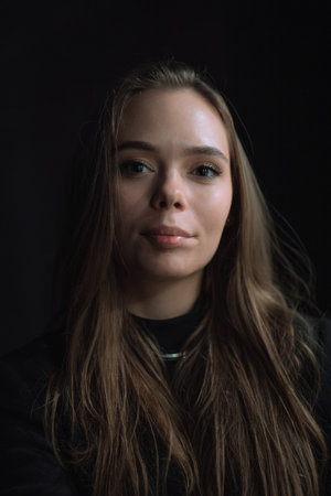 Front facing studio portrait of a young woman against a deep black background. Soft directional...