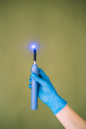 Close-up of a dental curing light with active LED tip held in a blue medical glove. Modern dental equipment isolated on green background, concept of dental treatment, hygiene, healthcare technology and professional dentistry tools.の写真素材