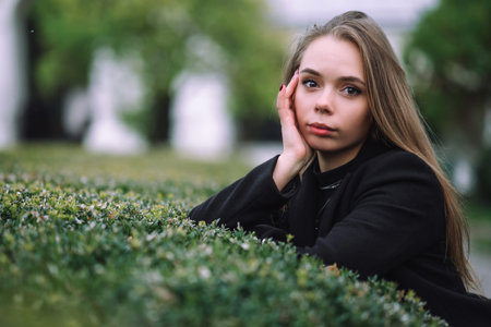A beautiful girl with long hair and a soft gaze leaning her chin on her hand over a trimmed green...