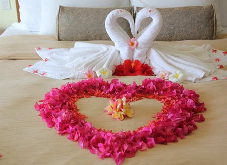 Close up of two white towel swans, with flowers shaped into a big heart on the bed of a honeymoon suite.の写真素材