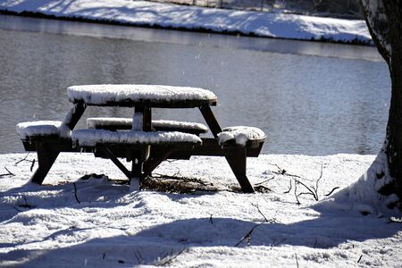 Snow covers the ground and a round wooden table with benches by the pondの写真素材
