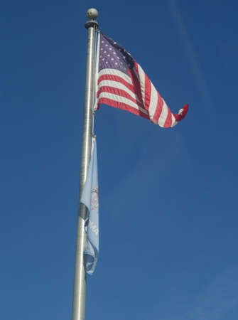 The United States flag waving in the air from a long pole, with the flag of Oklahoma State below