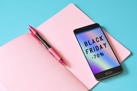 Smartphone with Black FRIDAY sale text on abstract holographic flow on display and pink notebook on blue table. Mock up for designの写真素材