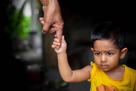 A baby boy confidently walks around holding on the finger of a senior old man. Family, Generation, Support and people concept. Dark background.の写真素材