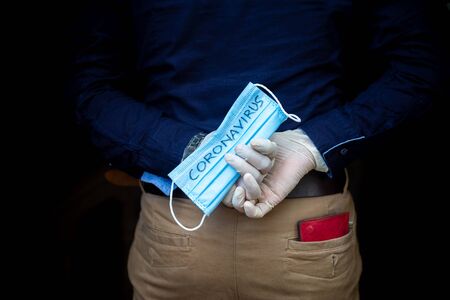 A man stands back holding a surgical mask with his hands wearing surgical gloves to protect against...