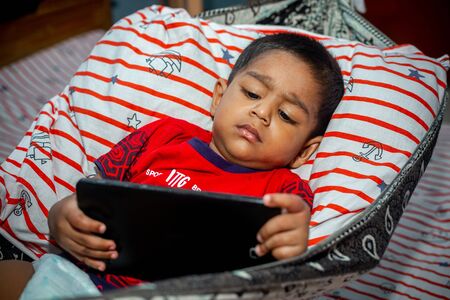 Before going to sleep a child is lying a homemade hammock and watching cartoons using a smartphone tab. Kids playing with smartphone. Mobile phone and internet addiction concept.の写真素材
