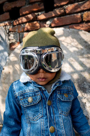 A pilot boy is wearing an old blue jeans jacket and hand gloves on a heavy winter day. A happy kid in a pilot costume and goggles. Close-up view on rustic damaged wall background.の写真素材