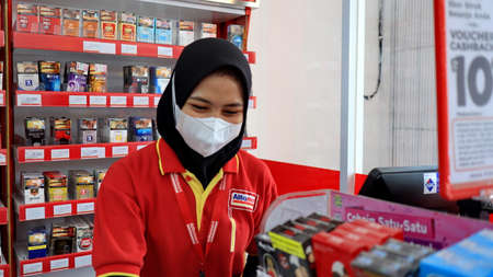 The female cashier of Alfamart is serving customers by using a face mask and according to health...
